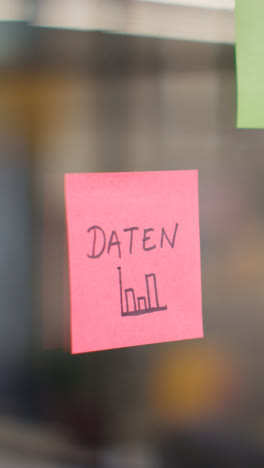 Vertical-Video-Close-Up-Of-Woman-Putting-Sticky-Note-With-German-Word-Daten-Or-Data-Written-On-It-Onto-Transparent-Screen-In-Office-